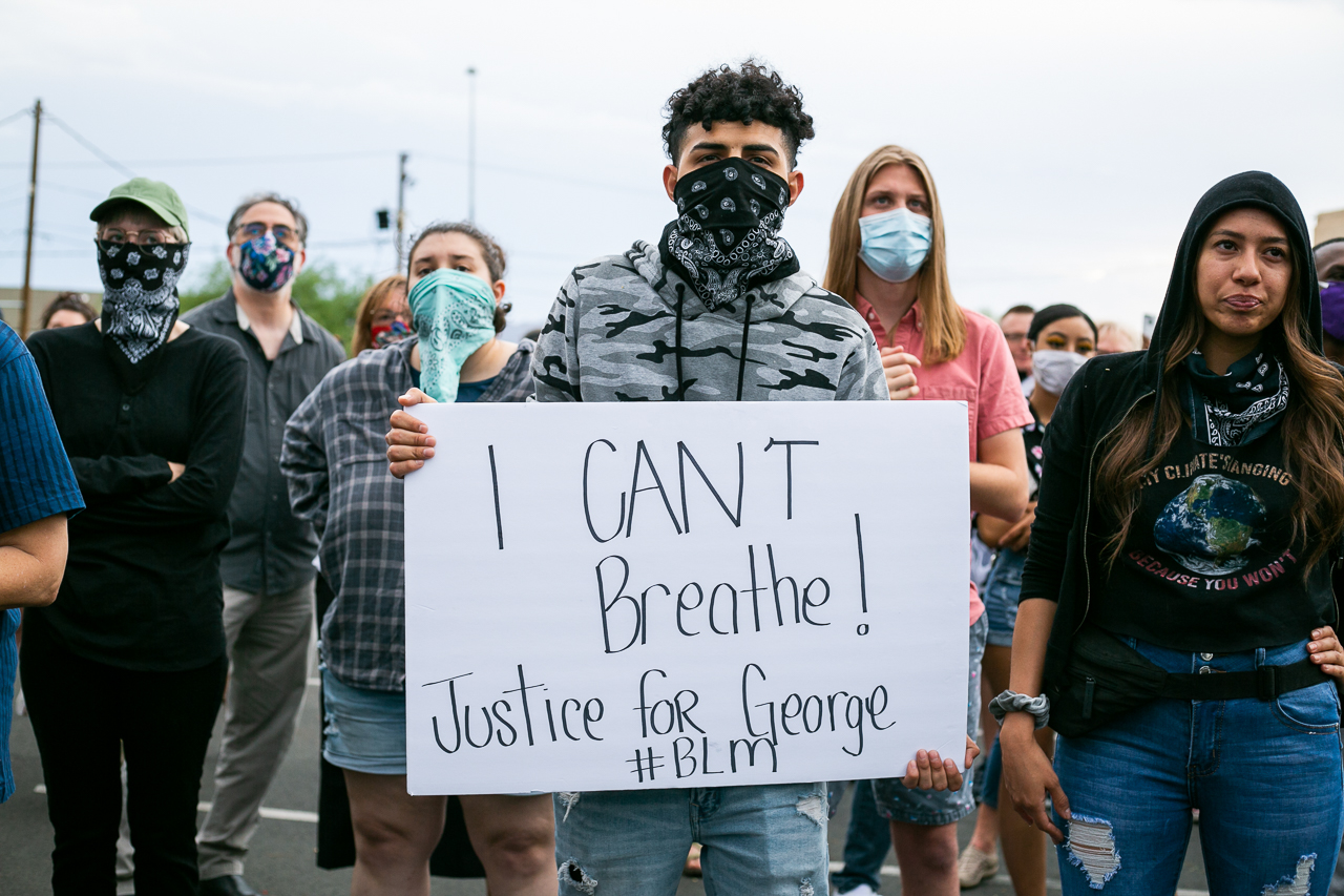 A young man with curly hair holds a sign that says "I can't Breath. Justice for George Floyd" with several people standing on either side of him.