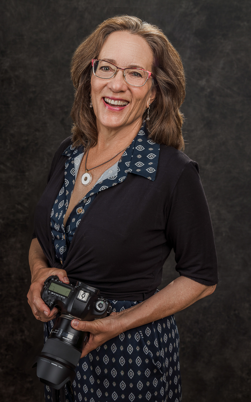 750x1200_WEB_MRP00186-Edit A white woman with shoulder-length hair, standing holding her camera in both hands. She is wearing a diamond-pattern jumpsuit withe a 3/4 sleeve black over wrap.