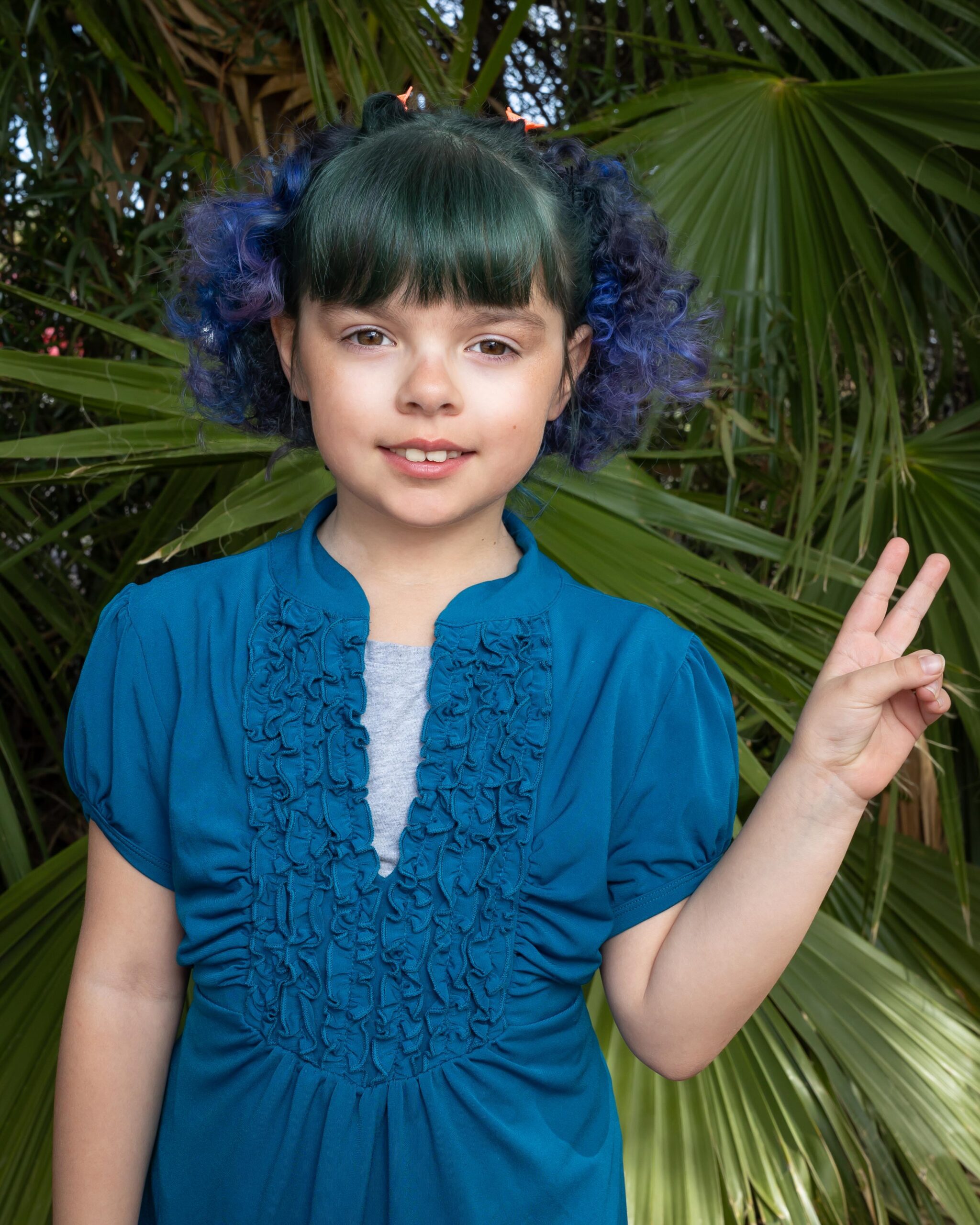 Young girl with blue hair, wearing a blue shirt and giving the peace sign during her elementary school portrait by Kathleen Dreier Photography