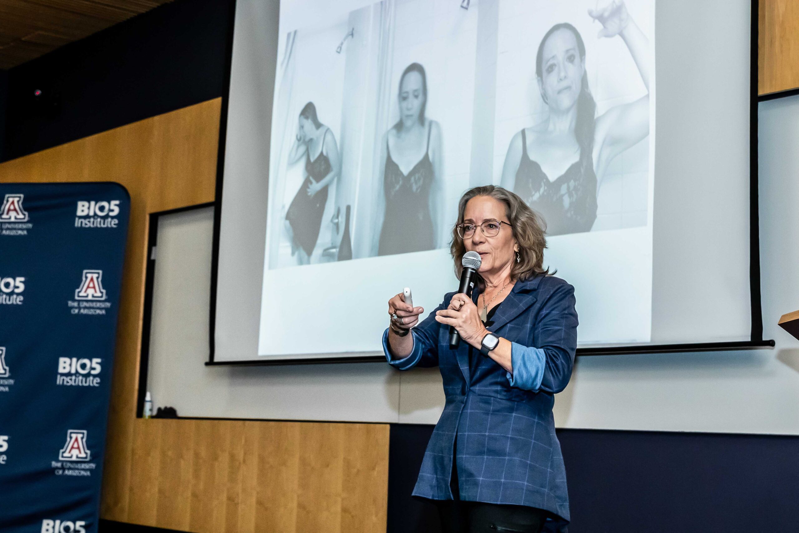 Creative Mornings – Kathleen Dreier “CYCLE” Kathleen Dreier wearing a blue jacket and holding a mic is giving a presentation in front of a projection screen with some of her photography images during her Creative Mornings presentation on the topic of Cycle.
